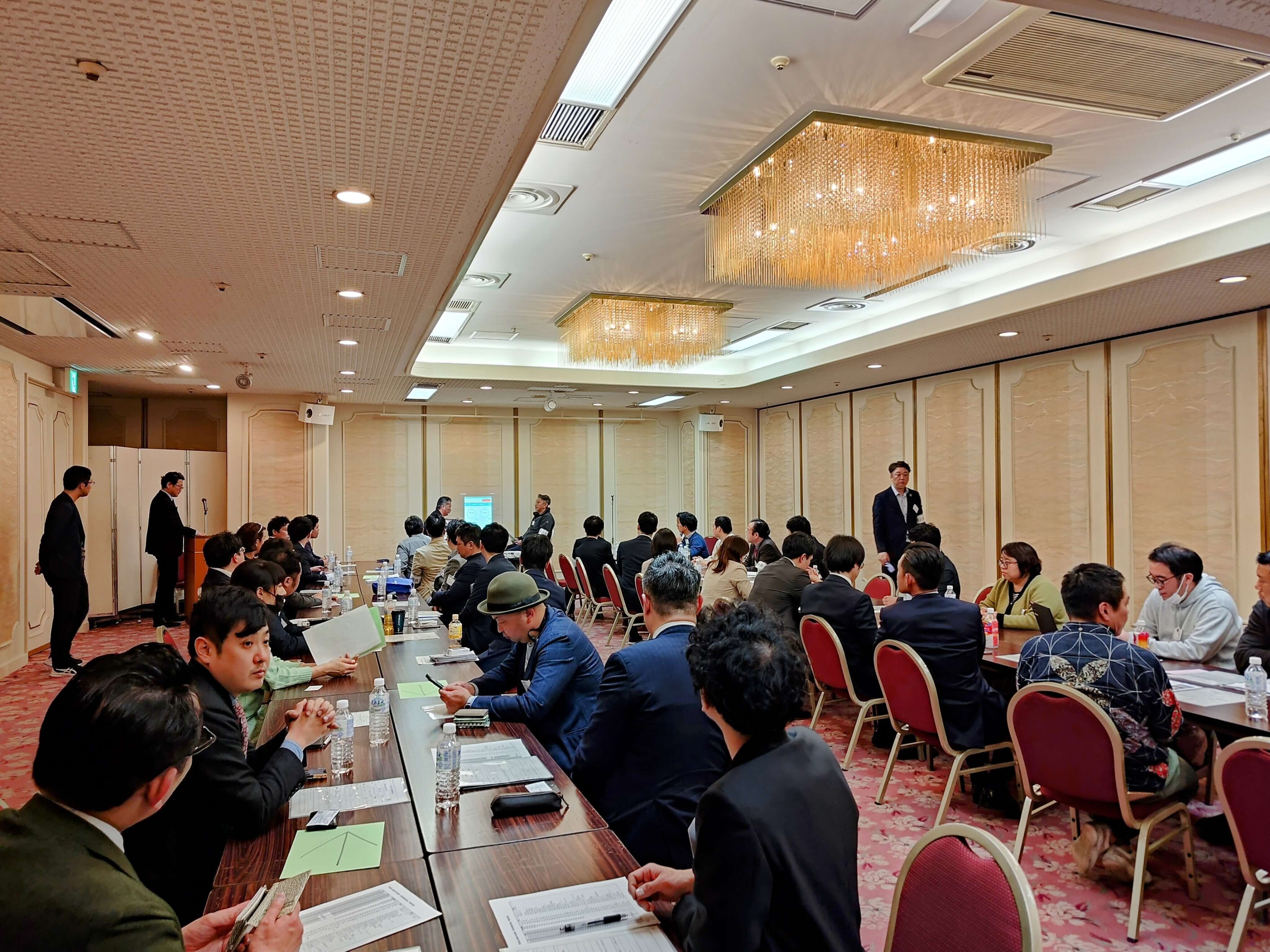 A roomful of business professionals seated at long tables, listening to presenters and engaging in networking activities at a formal business matching event in Tokyo.
