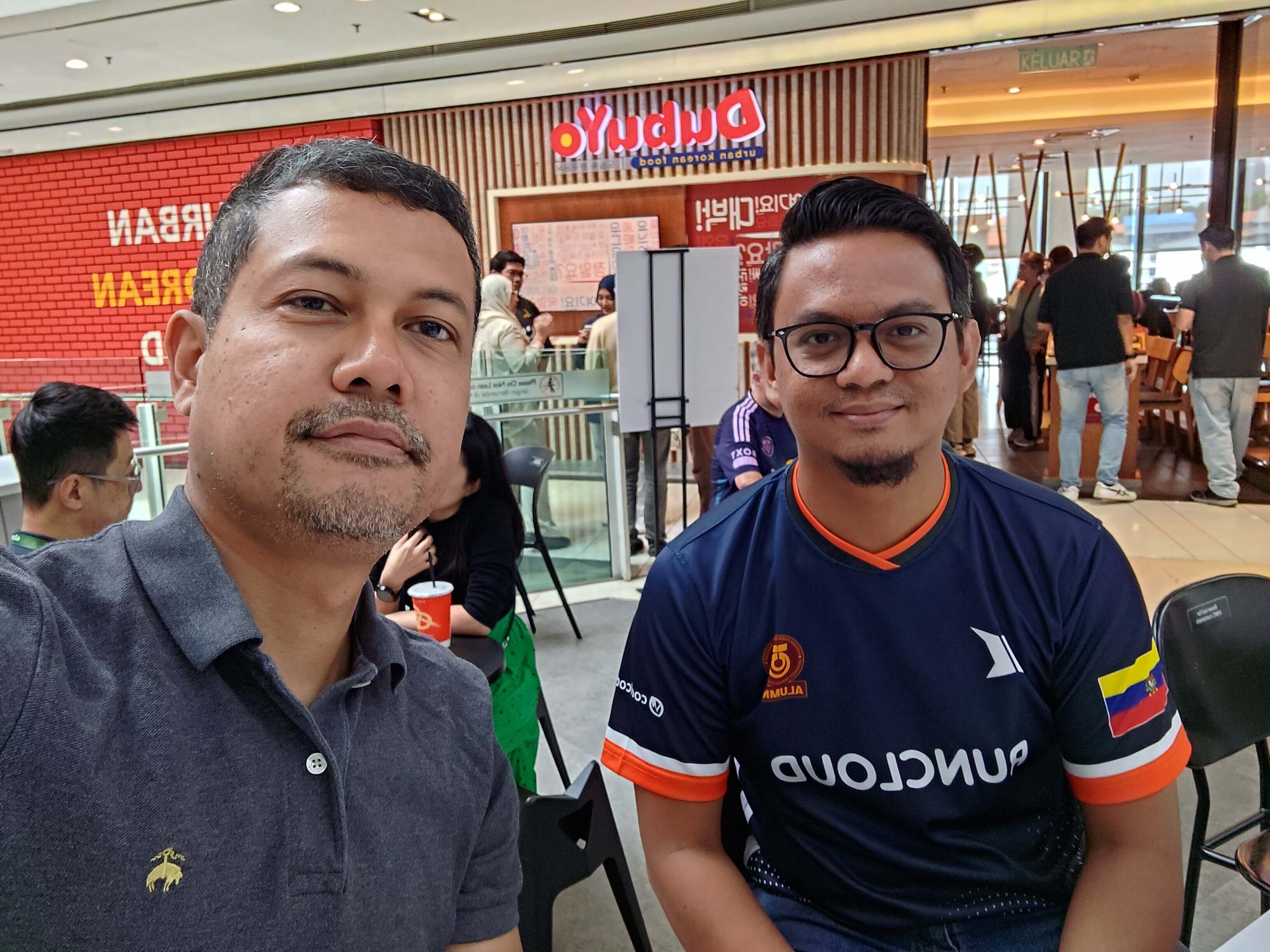 Two people posing for a wefie with a group of people in the background